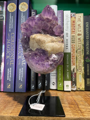 An amethyst cluster with calcite inclusion displayed on a metal stand, with a label in front, placed on a wooden surface with books in the background.